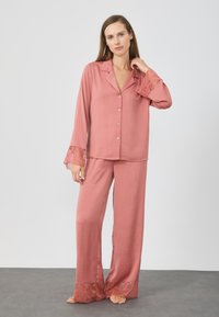Ružový saténový pyžamový set pozostávajúci z príjemne zapínacej blúzky s dlhým rukávom a širokých nohavíc, ktoré sú zdobené čipkou na manžetách a na okrajoch.