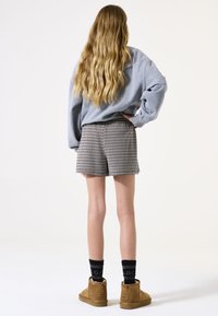 Sweat à capuche gris, shorts à carreaux, longueur genou, texture en mélange de laine. Bottines brunes, chaussettes noires avec logo. Cheveux raides, vue de dos.