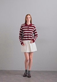 Maroon ja valkoinen raidallinen pitkähihainen polo-paita, jossa on napit, yhdistetty valkoiseen taitettuun hameeseen. Asusteina tummanharmaat sukat ja tummat loaferit.