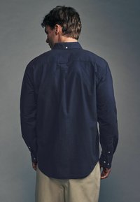 Námornícky modrá košeľa s dlhým rukávom, so záhybom na zadnej strane, s gombíkmi na manžetách a zaobleným lemom. Tkanina vyzerá hladko s jemným leskom.