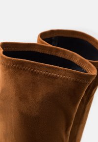Botas de tobillo de ante marrón con una textura suave, que cuentan con una abertura cosida y un forro interior negro. La forma es ligeramente caida.