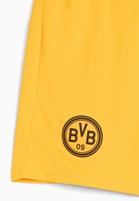Pantalones cortos deportivos amarillos con una tela texturizada. Presentan un logo negro circular con "BVB 09" en el centro, situado en el lado izquierdo.