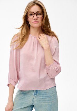 Kobieta o długich blond włosach, w okularach w kolorze żółwia, jasnoróżowej bluzce z podwiniętymi rękawami i jasnoniebieskich dżinsach, pozująca na białym tle.