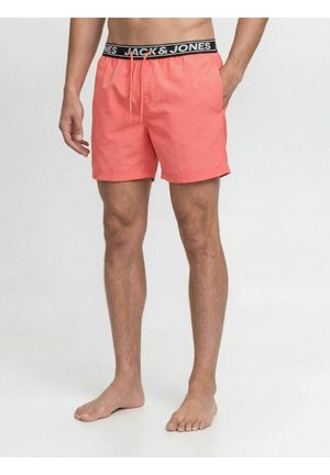 Mand iført koralfarvede badebukser med sort elastisk linning mærket "JACK & JONES", stående barfodet mod en ensfarvet baggrund.