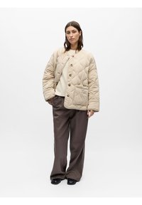 Γυναίκα με μπεζ μπουφάν καπιτονέ, κρεμ πουλόβερ, φαρδιά καφέ παντελόνια και μαύρα παπούτσια, που στέκεται μπροστά σε λευκό φόντο.