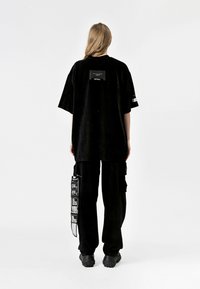 Camiseta oversize de pana negra con una etiqueta rectangular en la parte posterior, combinada con pantalones cargo negros que presentan texto blanco en una etiqueta colgante.