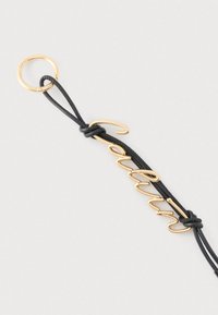 Χρυσό μπρελόκ Calvin προσαρτημένο σε μαύρο δερμάτινο κορδόνι με χρυστό σύρμα που σχηματίζει τη λέξη "Calvin" με καλλιγραφικά γράμματα.