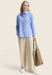 Camicia azzurro chiaro con bottoni, maniche lunghe, taglio ampio, abbinata a pantaloni beige a gamba larga e una borsa tote color cuoio. Look completato da sneaker bianche.