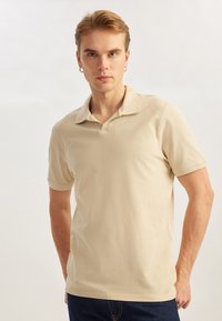Camisa polo de algodón beige con cuello clásico, mangas cortas y dobladillo recto, combinada con pantalones vaqueros azules, presentando un look casual.