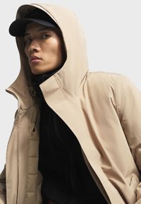 Lichtbeige, waterdichte jas met een capuchon. Voorzien van een rits- en knoopsluiting, elastische trekkoord en een zwarte binnenlaag.