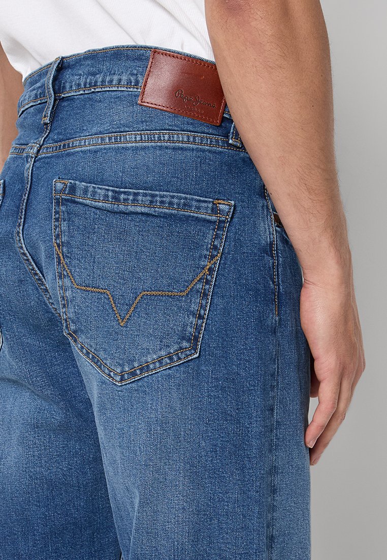 Zbliżenie na osobę noszącą niebieskie dżinsy Pepe Jeans, z widocznym przeszyciem tylnej kieszeni i brązową skórzaną metką na pasku.
