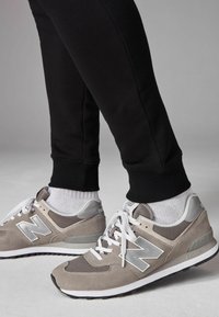Helle graue und beidge Wildledersneaker mit einem Mesh-Obermaterial, weißen Schnürsenkeln, umrandetem Logo und schwarzer Gummisohle, kombiniert mit schwarzen umgekrempelten Jogginghosen.