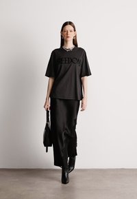 Μαύρο βαμβακερό t-shirt με τη λέξη "ΕΛΕΥΘΕΡΙΑ" εκτυπωμένη με έντονα γράμματα, συνδυασμένο με μαύρη σατέν φούστα και μαύρες μπότες με τετράγωνο τακούνι. Το μοντέλο κρατά μια μαύρη τσάντα.
