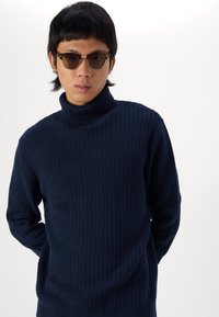 Jersey de punto azul marino con un alto cuello acanalado y patrón texturizado de zigzag; la modelo lleva gafas de sol redondas y está de pie con las manos detrás de la espalda.