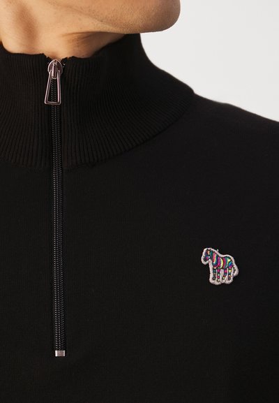 Fekete kötött pulóver magas gallérral és cipzárral, a bal mellen egy kis, színes hímzett zebra díszíti.