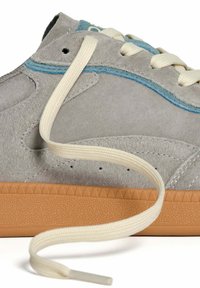 Sneaker grigia in suede con bordi blu, suola in gomma e lacci color crema che si arricciano davanti su uno sfondo bianco.