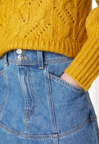 Pull en tricot jaune avec un motif texturé, associé à un jean denim bleu taille haute avec des boutons métalliques et des poches latérales.