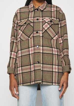 Vrouw draagt een olijfgroen en roze geruit blouse met knopen en twee borstzakken, gecombineerd met een lichtblauwe spijkerbroek, staand tegen een witte achtergrond.