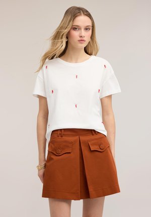 Jeune femme aux longs cheveux blonds portant un t-shirt blanc avec des accents rouges et une jupe marron avec de grandes poches boutonnées.