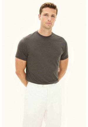 Giovane uomo con capelli castani corti che indossa una maglietta grigio scuro e pantaloni bianchi, in piedi con le mani dietro la schiena su uno sfondo chiaro.