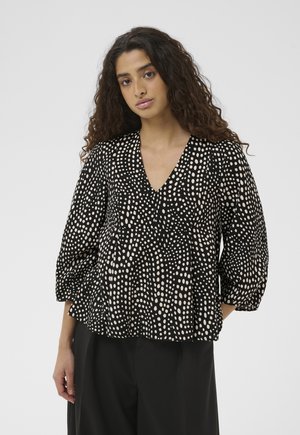 Woman with long curly hair wearing a black blouse with white irregular polka dots and loose black pants against plain background.