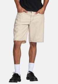 Shorts in denim beige con due tasche frontali, taglio dritto e dettagli cuciti. Indossati con una maglietta nera, calzini bianchi e scarpe nere.