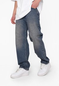 Jeans in denim blu con una vestibilità comoda, texture sbiadita e cuciture visibili. Abbinati a sneakers bianche, con un classico design con lacci.