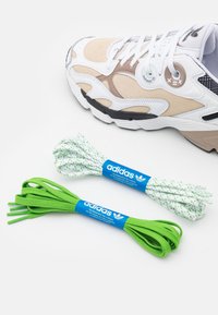 Adidas-skor i vitt, beige och svart med texturerat mocka och mesh-detaljer. Inkluderar två par skosnören: ett grönt, ett vitt med gröna accenter.