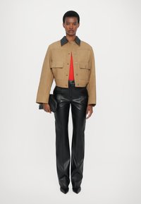Jachetă scurtă bej cu guler negru, două buzunare frontale, asortată cu pantaloni din piele negri și o poșetă neagră. Modelul stă pe un fundal alb.