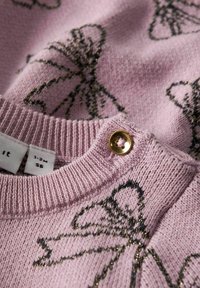 Maglione rosa lavorato a maglia con motivi a fiocco neri, caratterizzato da un colletto a costine testurizzato e un bottone dorato vicino al colletto. Etichetta della taglia visibile.