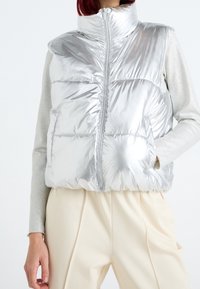 Silberne Puffweste mit hohem Kragen, Reißverschluss und horizontalen Steppnähten; getragen über einem hellgrauen Langarmshirt und cremefarbenen Hosen.