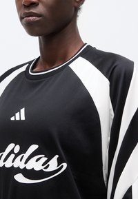 Κοντινό πλάνο ενός ατόμου που φοράει αθλητική φανέλα Adidas σε μαύρο και άσπρο, με εμφανές το στήθος και μέρος του προσώπου, μπροστά σε μονόχρωμο φόντο.