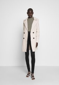 Kaksirivinen, vaaleanbeige ruudullinen takki, jossa on leveä kaulus, mustat istuvat farkut ja valkoiset nilkkaketjukenkiä, taustalla yksinkertainen tausta.