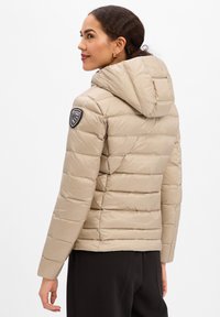 Donna di profilo che indossa una giacca trapuntata beige con cappuccio e una toppa con logo sulla manica sinistra, e pantaloni neri, su sfondo bianco.