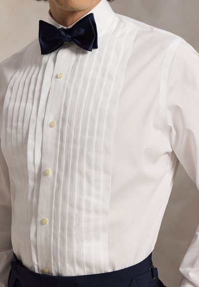 Uomo che indossa una camicia bianca plissettata con bottoni crema, papillon nero e pantaloni scuri, mostrato da metà petto alla vita.