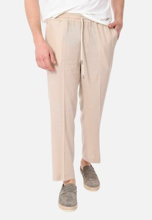 Uomo con pantaloni beige con coulisse e scarpe slip-on grigie, mani in tasca, in piedi su uno sfondo bianco semplice.