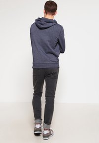 Sudadera azul marino con capucha y cordones, puños acanalados, combinada con jeans negros. Zapatillas gris y blanca con acentos verdes.