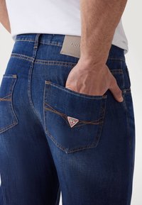 Traperice tamnoplave boje s narančastim šavovima, koje imaju trokutasti "Guess" logo na stražnjem džepu i sivi etiketu na struku.