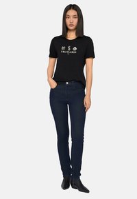 Camiseta de algodón negra con un logo y detalles gráficos en el frente, combinada con jeans azul oscuro y botas de tobillo negras. Diseño simple y ajustado.