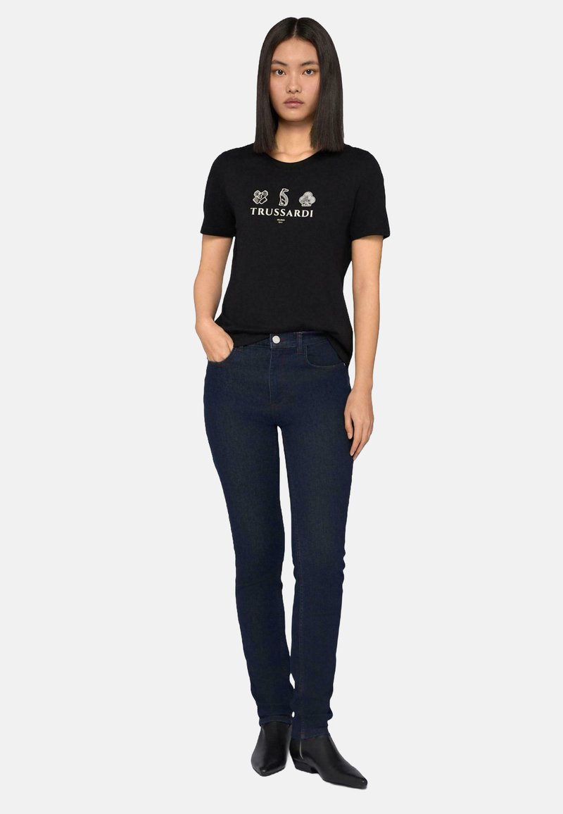 Camiseta de algodón negra con un logo y detalles gráficos en el frente, combinada con jeans azul oscuro y botas de tobillo negras. Diseño simple y ajustado.