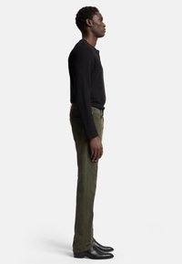 Maglietta nera a maniche lunghe, pantaloni slim-fit verde oliva e stivali neri alla caviglia. Design semplice, texture liscia e vestibilità sartoriale in evidenza.