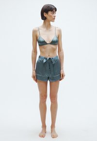 Bralette verde in satin con spalline sottili e pantaloncini a vita alta con fantasia. I pantaloncini hanno un laccetto frontale e una vestibilità comoda. Piedi nudi.