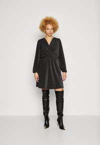 Μαύρο γκλιτερένιο φόρεμα με σχέδιο τύπου περιτυλίγματος, μακριά φουσκωτά μανίκια και ζωνάκι στη μέση. Συνδυασμένο με ψηλές, μαύρες δερμάτινες μπότες με μυτερές άκρες.