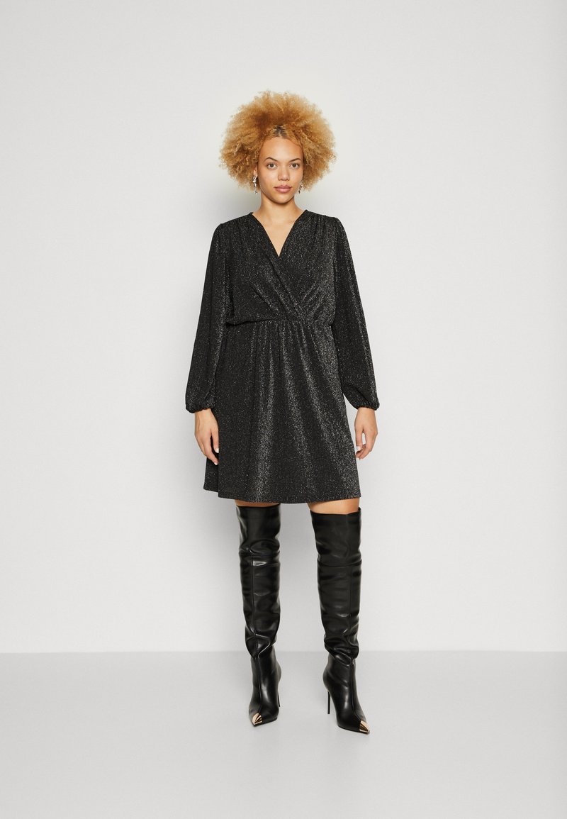 Μαύρο γκλιτερένιο φόρεμα με σχέδιο τύπου περιτυλίγματος, μακριά φουσκωτά μανίκια και ζωνάκι στη μέση. Συνδυασμένο με ψηλές, μαύρες δερμάτινες μπότες με μυτερές άκρες.