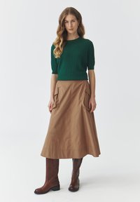 Groene ribgebreide trui gecombineerd met een tan A-lijn rok met grote zijzakken. Model draagt bruine kniehoge laarzen.