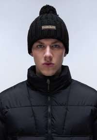 Black knit beanie with a pom-pom on top and a logo patch. Worn with a black puffer jacket featuring a high collar and zip closure.