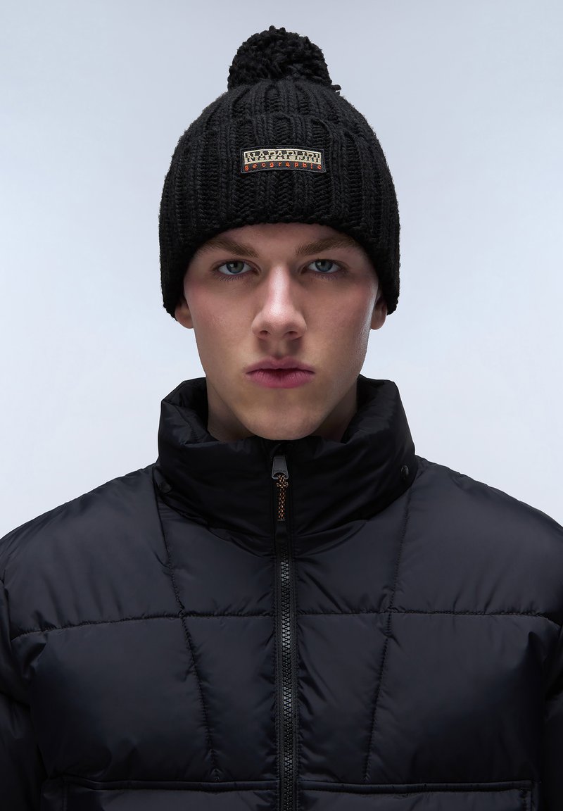 Black knit beanie with a pom-pom on top and a logo patch. Worn with a black puffer jacket featuring a high collar and zip closure.