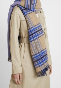 Personne portant un trench-coat beige avec une large écharpe à carreaux bleu et beige enroulée autour du cou, pendante à l'avant avec des extrémités à franges.