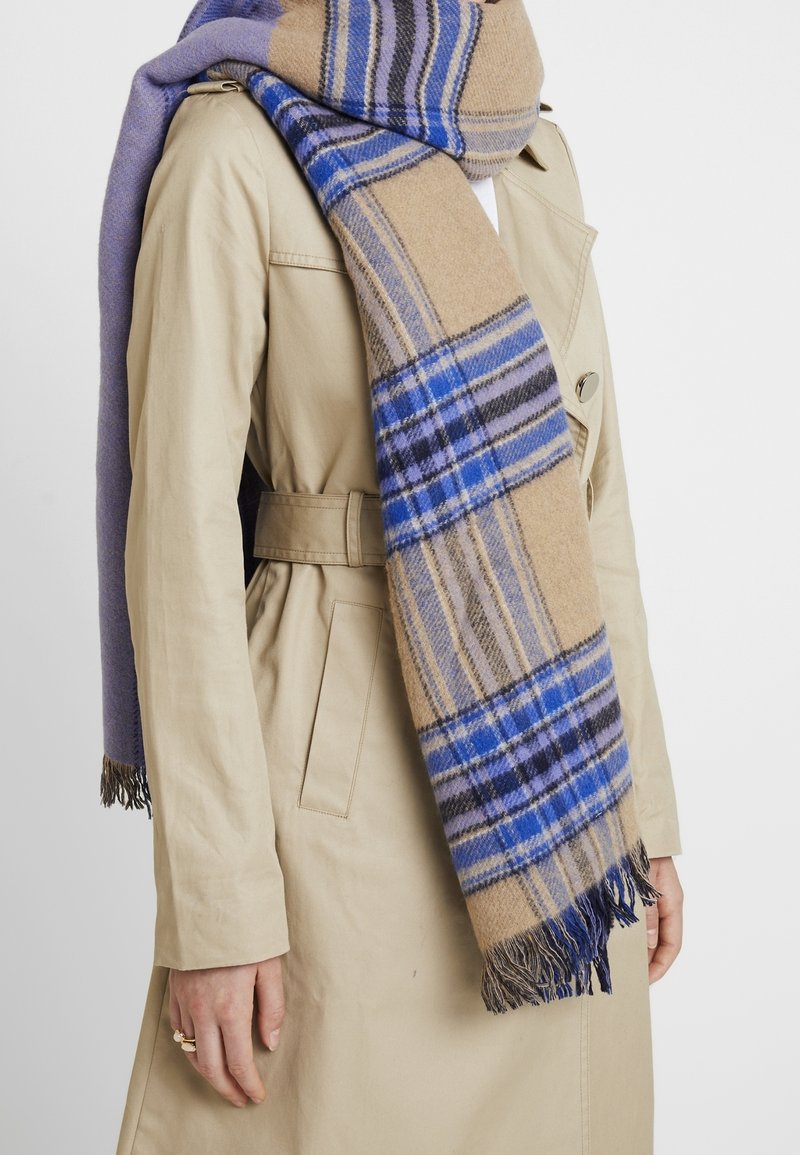 Personne portant un trench-coat beige avec une large écharpe à carreaux bleu et beige enroulée autour du cou, pendante à l'avant avec des extrémités à franges.
