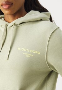 Hoody verde claro com bolso frontal, capuz com cordão e texto amarelo: "BJÖRN BORG STOCKHOLM 1956" no peito. Textura de tecido suave.